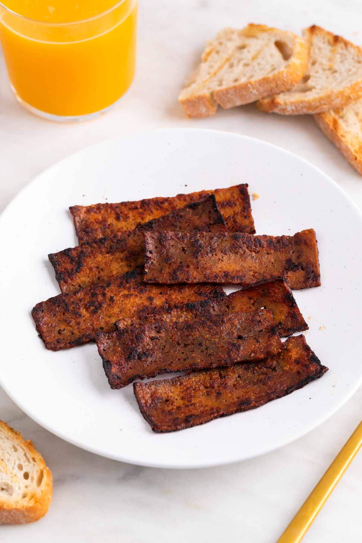 Tofu bacon on a plate with bread and orange juice around it.