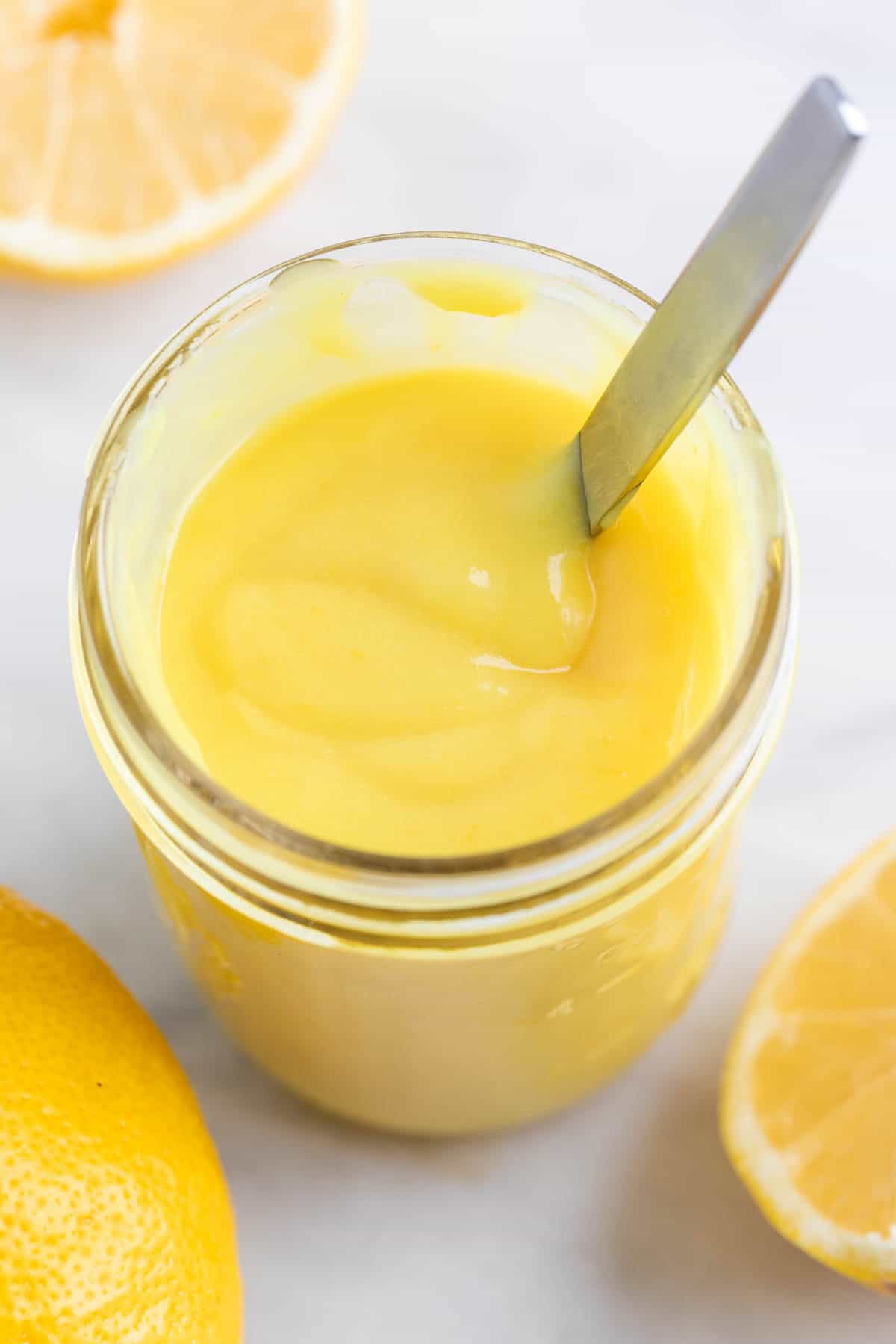 Jar of vegan lemon curd with a spoon inside and lemons around.