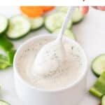 A spoon dipped into a bowl of vegan ranch dressing, surrounded by crudités.