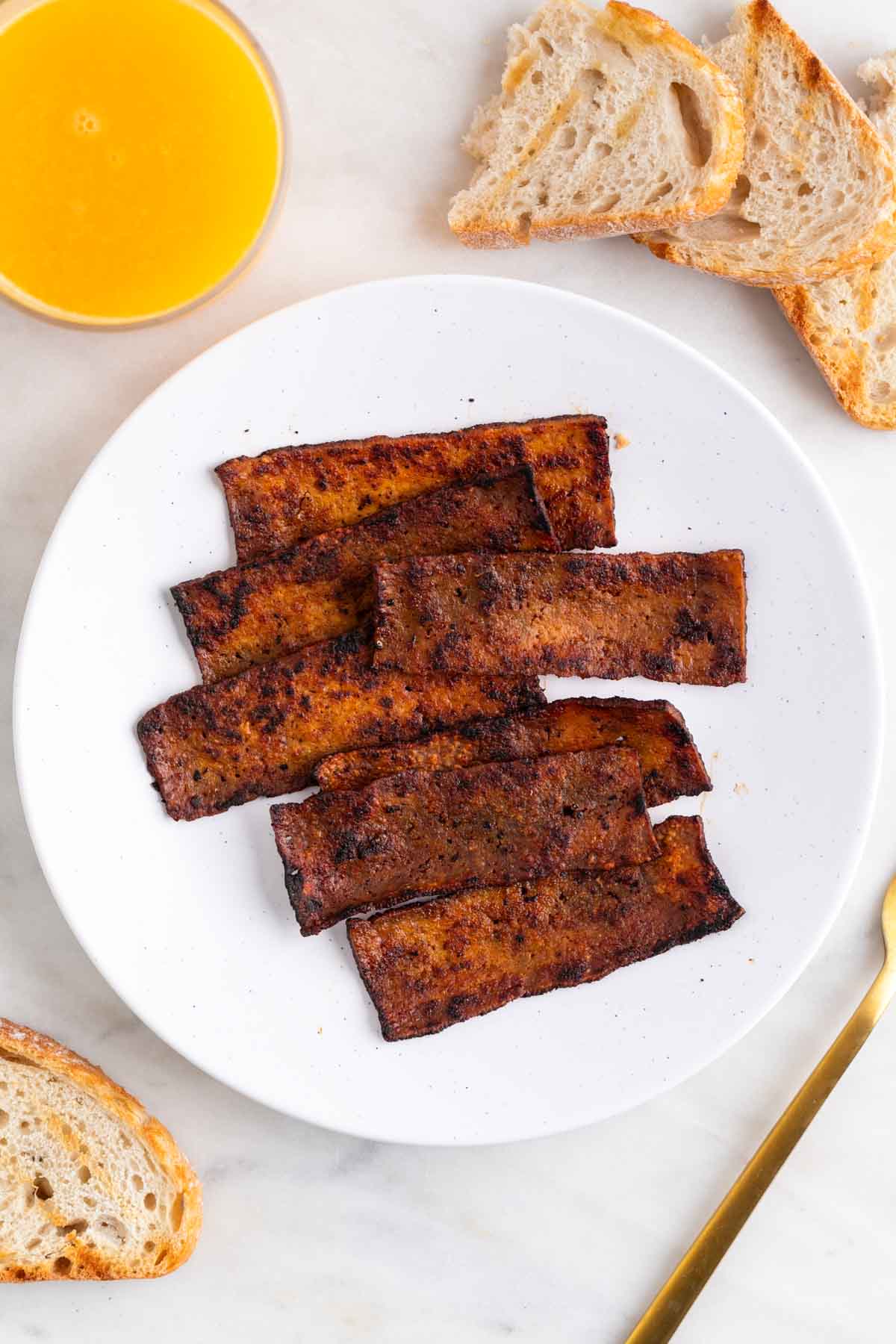 Tofu bacon served on a plate with bread and a glass of orange juice.