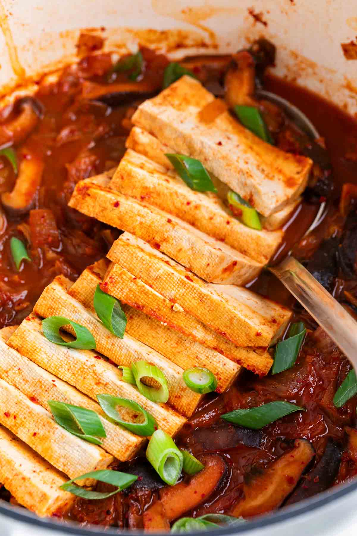 Vegan kimchi stew in a large pot, topped with scallions and a ladle inside.