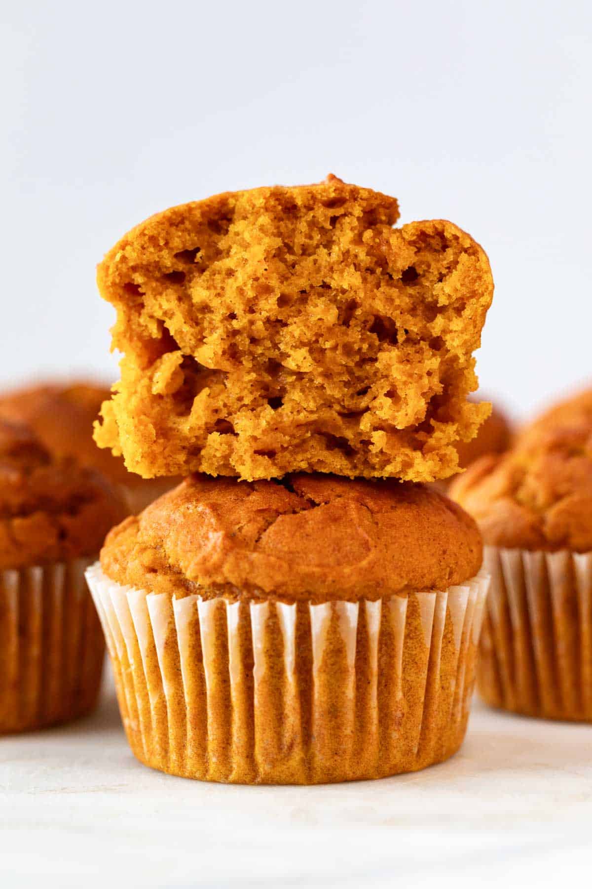 Stacked vegan pumpkin muffins with the top one sliced, surrounded by more muffins.