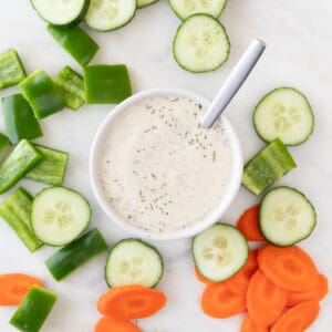 A spoon rests in a bowl of vegan ranch dressing, surrounded by crudités.