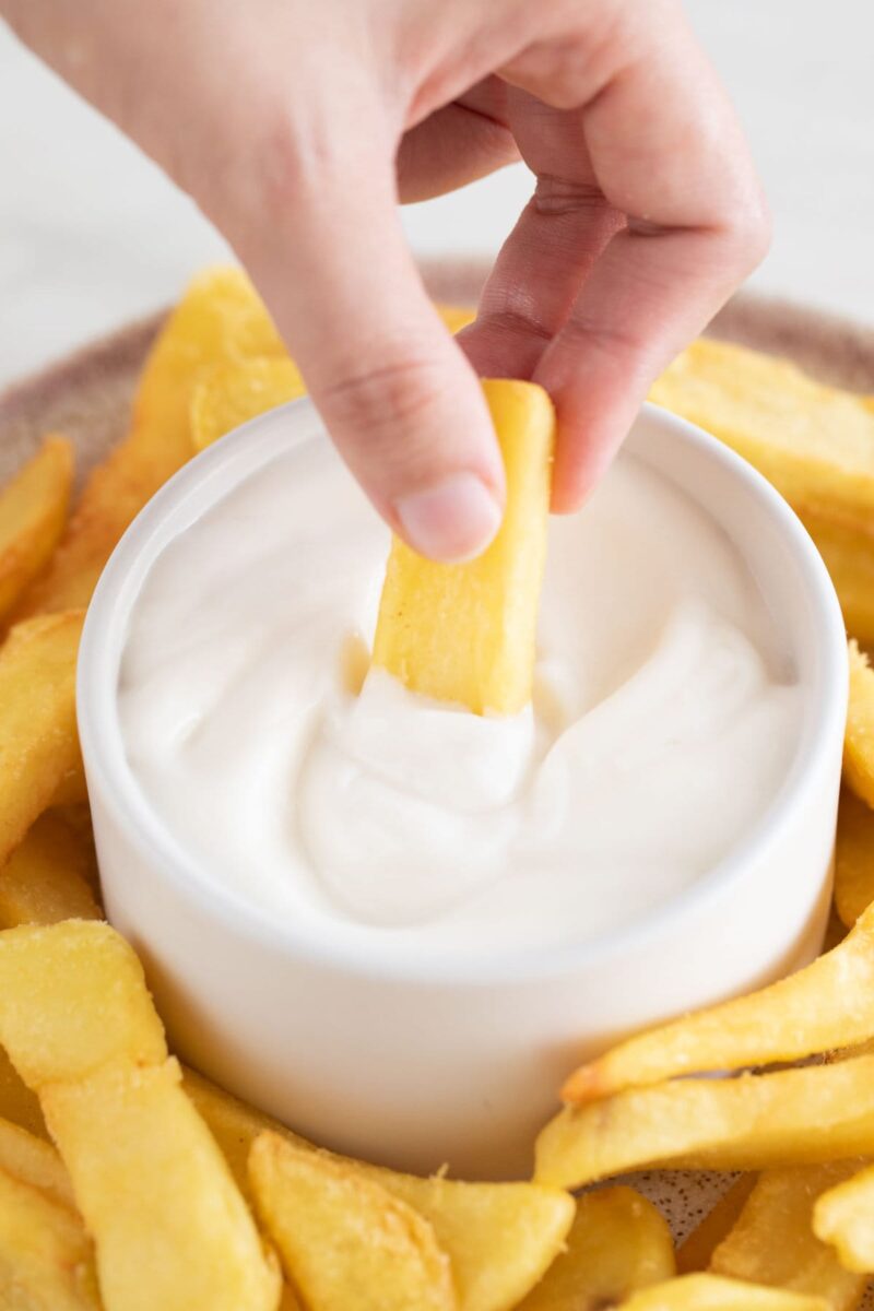 Photo of a dish with French fries and a small bowl with vegan aioli and a hand dipping a fry into the sauce.