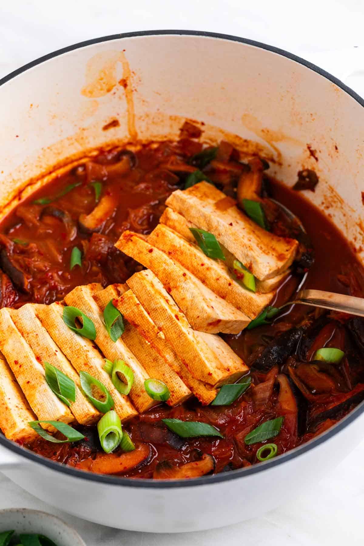 Vegan kimchi stew in a large pot with scallions on top and a ladle inside.