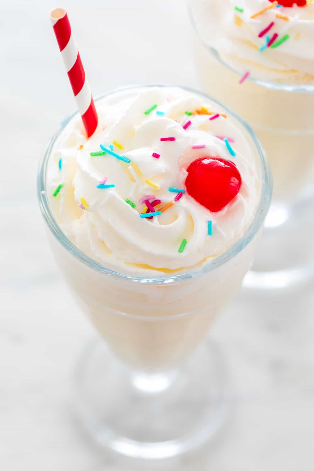 Vegan milkshake in a glass, topped with whipped cream, sprinkles, a cherry, and a straw.