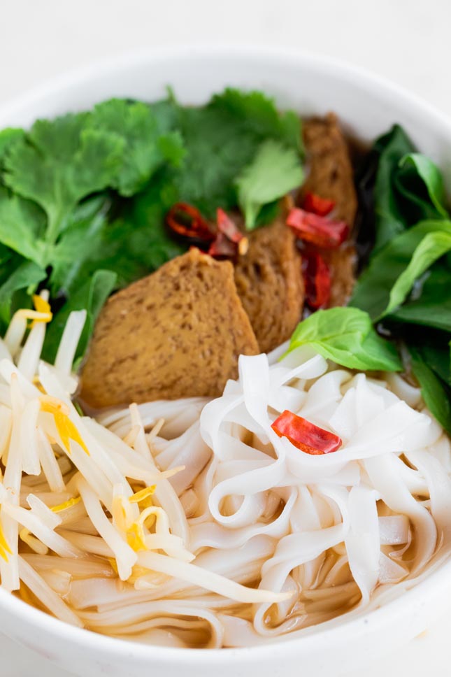 A close shot of a bowl with vegan pho topped with seitan, noodles, cilantro, basil and chili flakes
