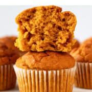 Stacked vegan pumpkin muffins with the top one sliced, surrounded by more muffins.