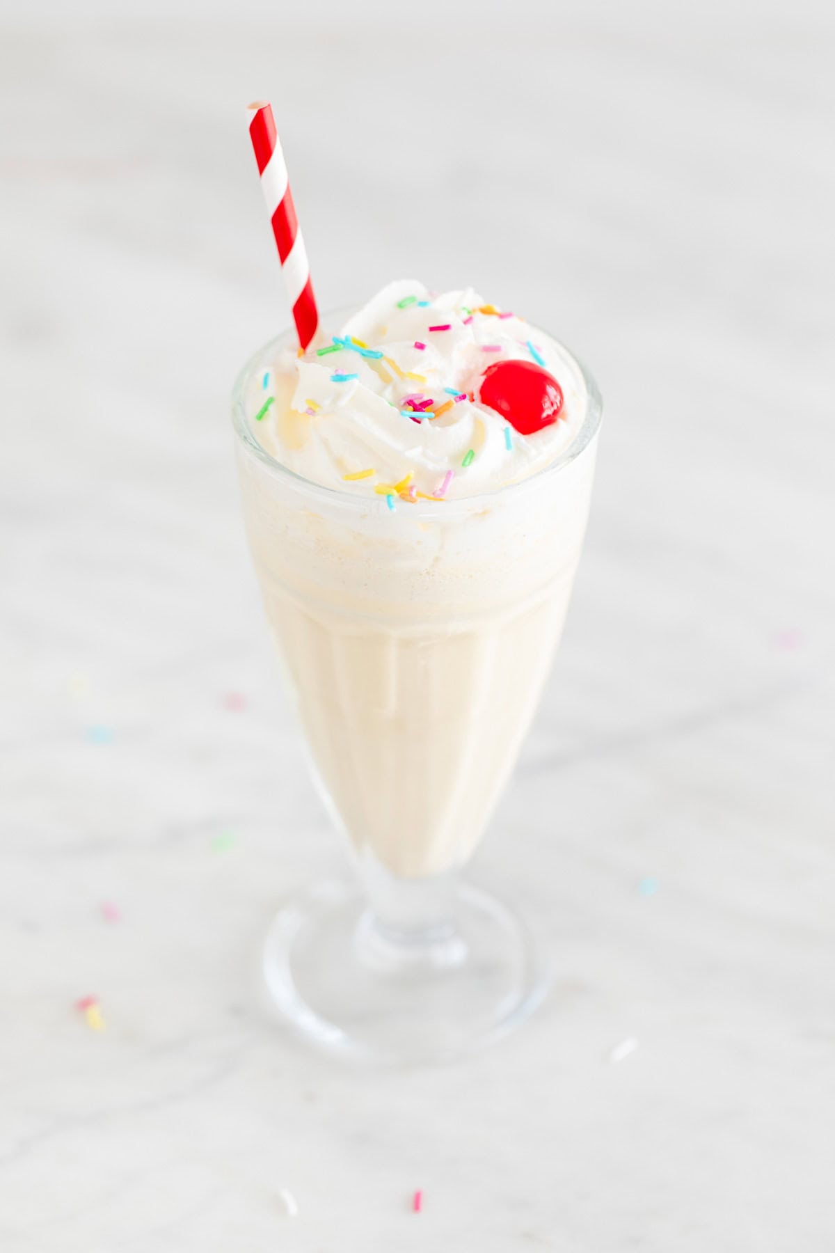 Glass of vegan milkshake topped with vegan whipped cream, colorful sprinkles, and a cherry.
